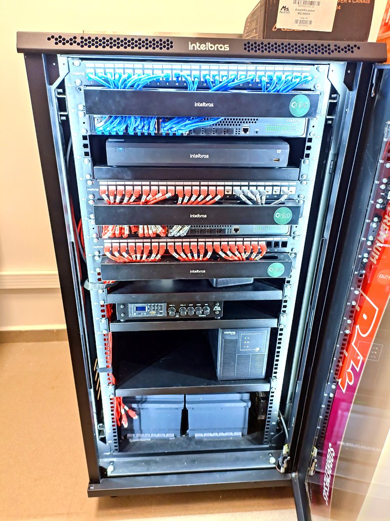 Rack de rede aberto com patch panels, switches Intelbras e cabeamento organizado por cores, identificado pela S2D.